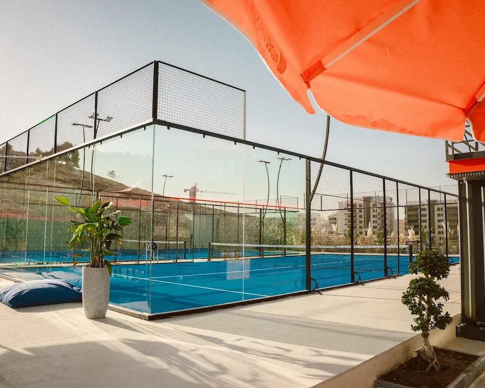 A blue Padel Mania court behind glass walls, seen from the shaded terrace in daylight