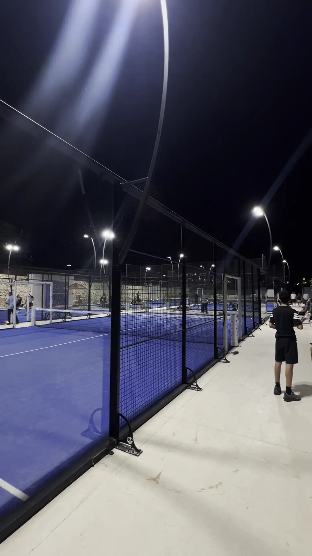 Floodlit blue padel courts behind the fence at Padel Mania at night